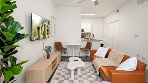 A modern living room with a sofa, coffee table, and TV. There is a dining area with chairs and a table. The room has white walls and a ceiling fan. - Modern Tempe 1BR • AC + Pool + Gym • 5 Min to ASU (Tempe)