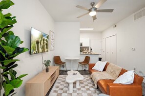 Ein modernes Wohnzimmer mit Sofa, Couchtisch und Fernseher. Es gibt eine Essecke mit Stühlen und Tisch. Der Raum hat weiße Wände und einen Deckenventilator.