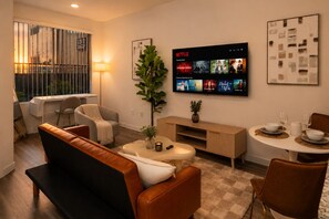 Ein modernes Wohnzimmer mit einem Flachbildfernseher, der Netflix-Inhalte zeigt, einem Essbereich mit rundem Tisch und Stühlen sowie einer Sitzecke mit einer Couch und Sesseln.