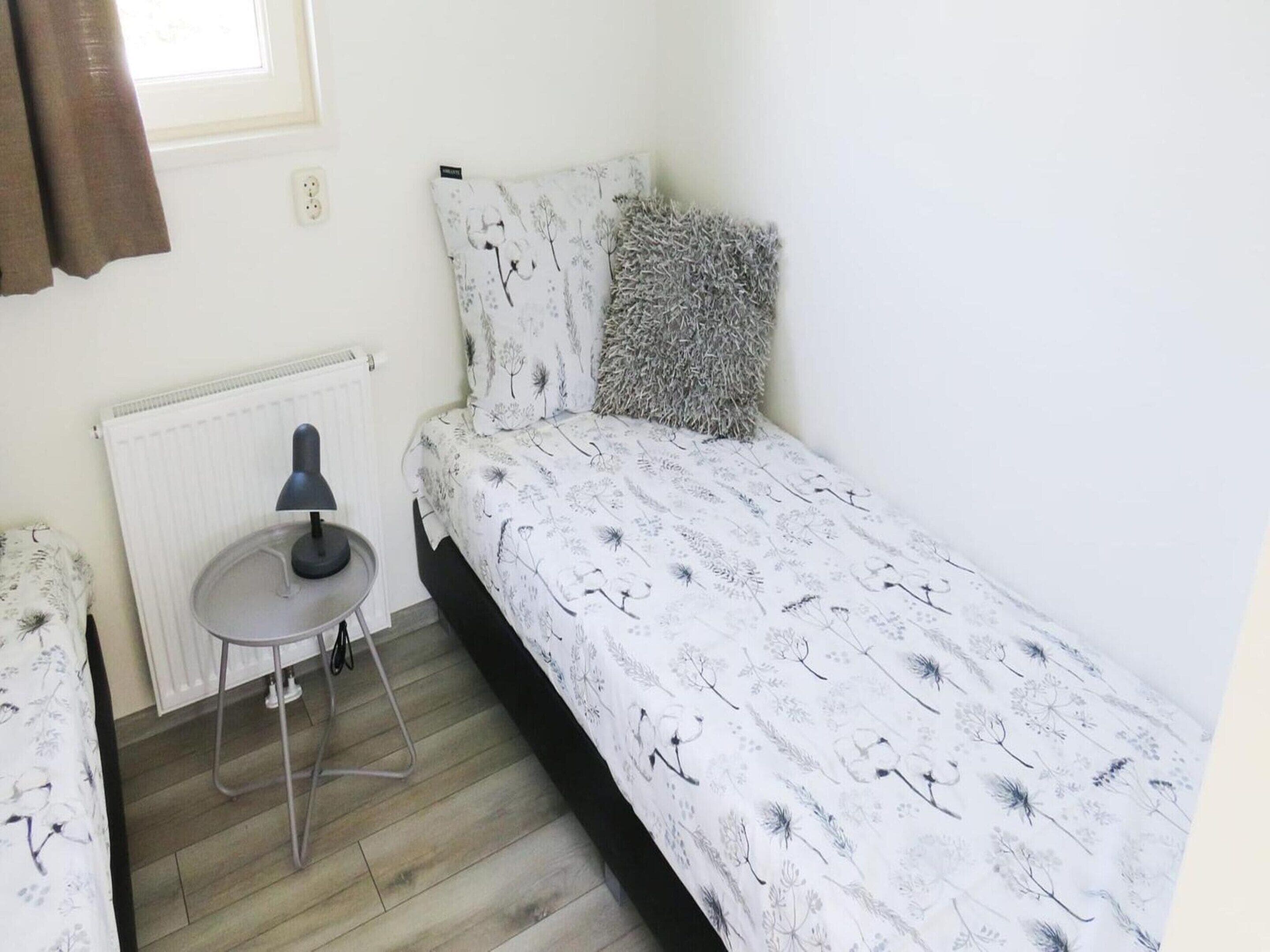 A small, single-bed room with a radiator, a table with a lamp, and a window with curtains.