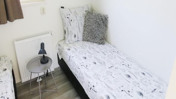 A small, single-bed room with a radiator, a table with a lamp, and a window with curtains.