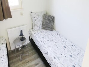 Una habitación pequeña con cama individual, un radiador, una mesa con lámpara y una ventana con cortinas.