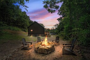 A rustic outdoor seating area with a stone fire pit and Adirondack chairs, surrounded by a wooden cabin and dense forest. - Sommerhus @ Skyline Drive | Modern Scandi Cabin on 6 Acres | 2BR, Sleeps 4 (Sperryville)