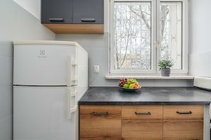 A kitchen with a white refrigerator, wooden cabinets, a black countertop, a window with a view of trees, and a bowl of fruit on the windowsill. - Family Stay on Boguckiego (Warsaw)