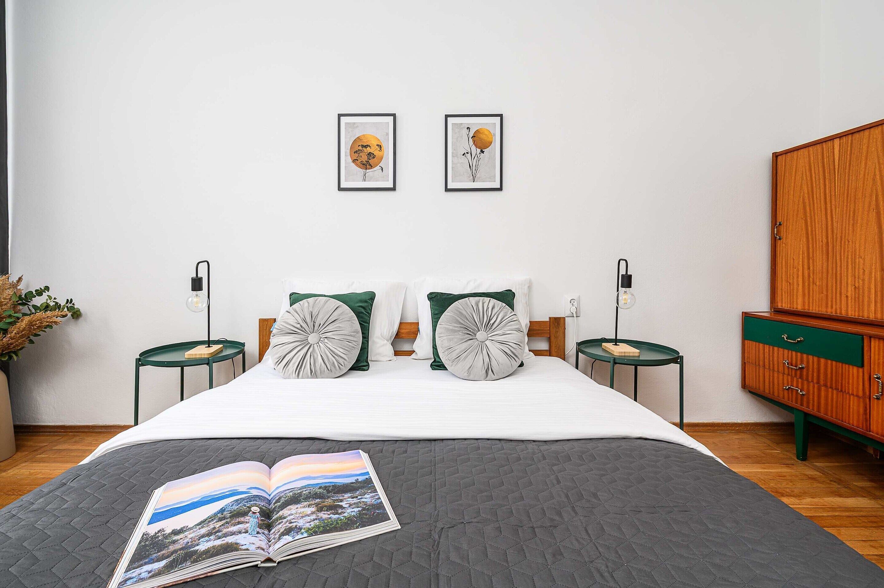 A neatly arranged bedroom with a bed, two bedside tables, a nightstand, a wardrobe, and two framed artworks on the wall.