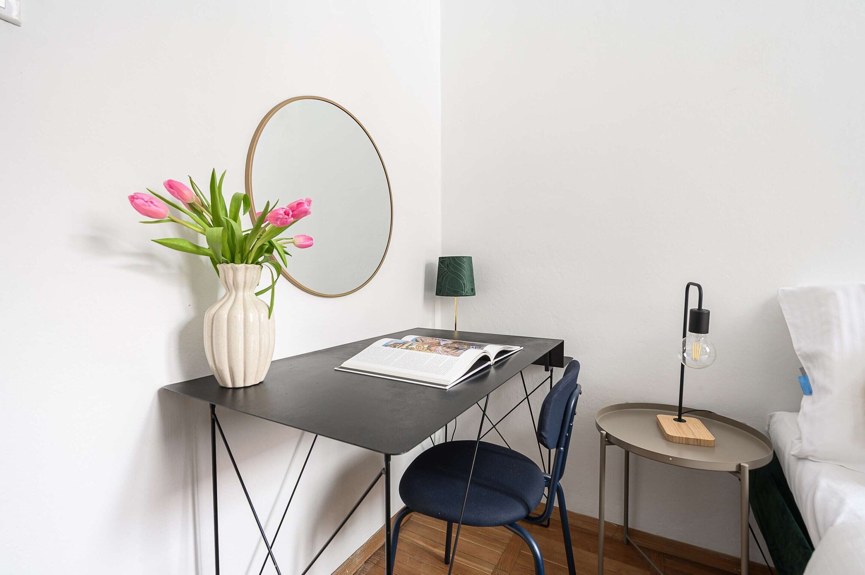 Una habitación moderna con escritorio, silla, espejo redondo, una pequeña mesa con lámpara y cama.