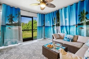 A room with a view of a tropical beach, a ceiling fan, and a sofa with decorative pillows. - Wellness Retreat 2 Min to Warm Mineral Springs (North Port)