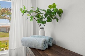 Ein Fenster mit weißen Vorhängen, eine weiße Vase mit grünen Blättern und zwei aufgerollte Handtücher auf einem Holzbrett.