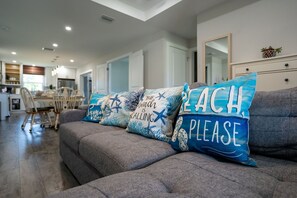 A modern living room with a grey sofa, a dining area in the background, and decorative pillows with beach-themed designs. - Wellness Retreat 2 Min to Warm Mineral Springs (North Port)