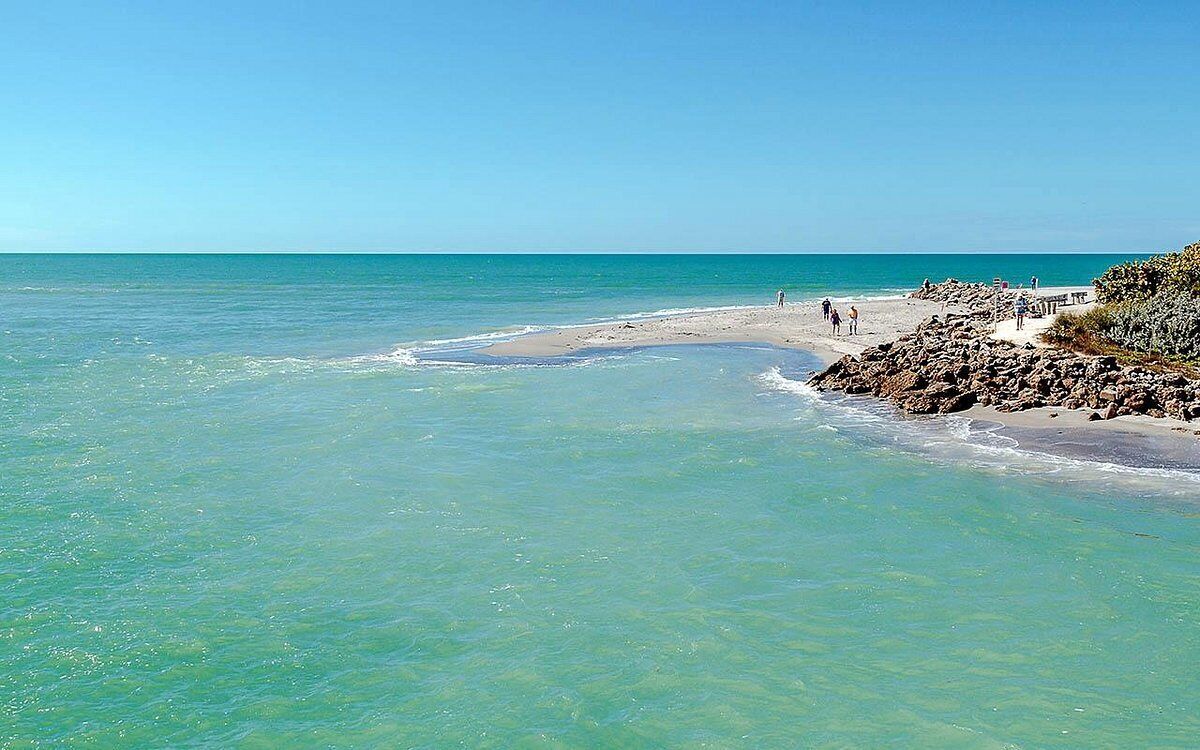 Una spiaggia con acque cristalline turchesi, costa rocciosa e alcune persone che camminano lungo il sentiero.