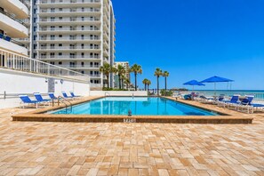A swimming pool with lounge chairs and umbrellas, surrounded by a brick deck and a high-rise building. - Ocean/River View | Heated Pool | Royal Atlantic 8D (Daytona Beach)
