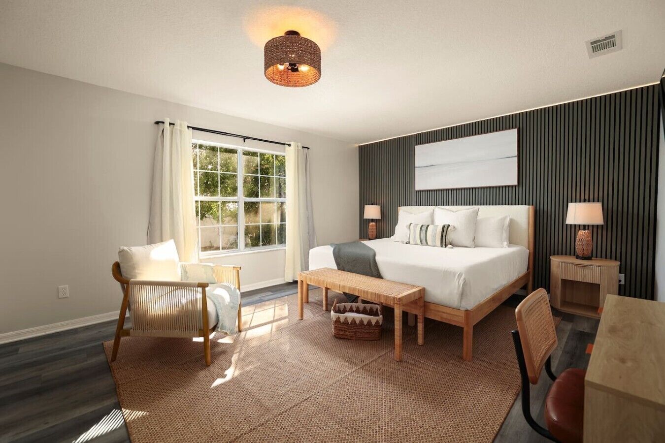 A hotel room with a bed, a desk, a chair, a window, and a wall-mounted lamp.