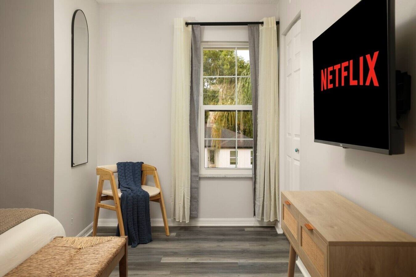Habitación de hotel con cama, una silla de madera, una mesita, televisor montado en la pared y una ventana con cortinas.