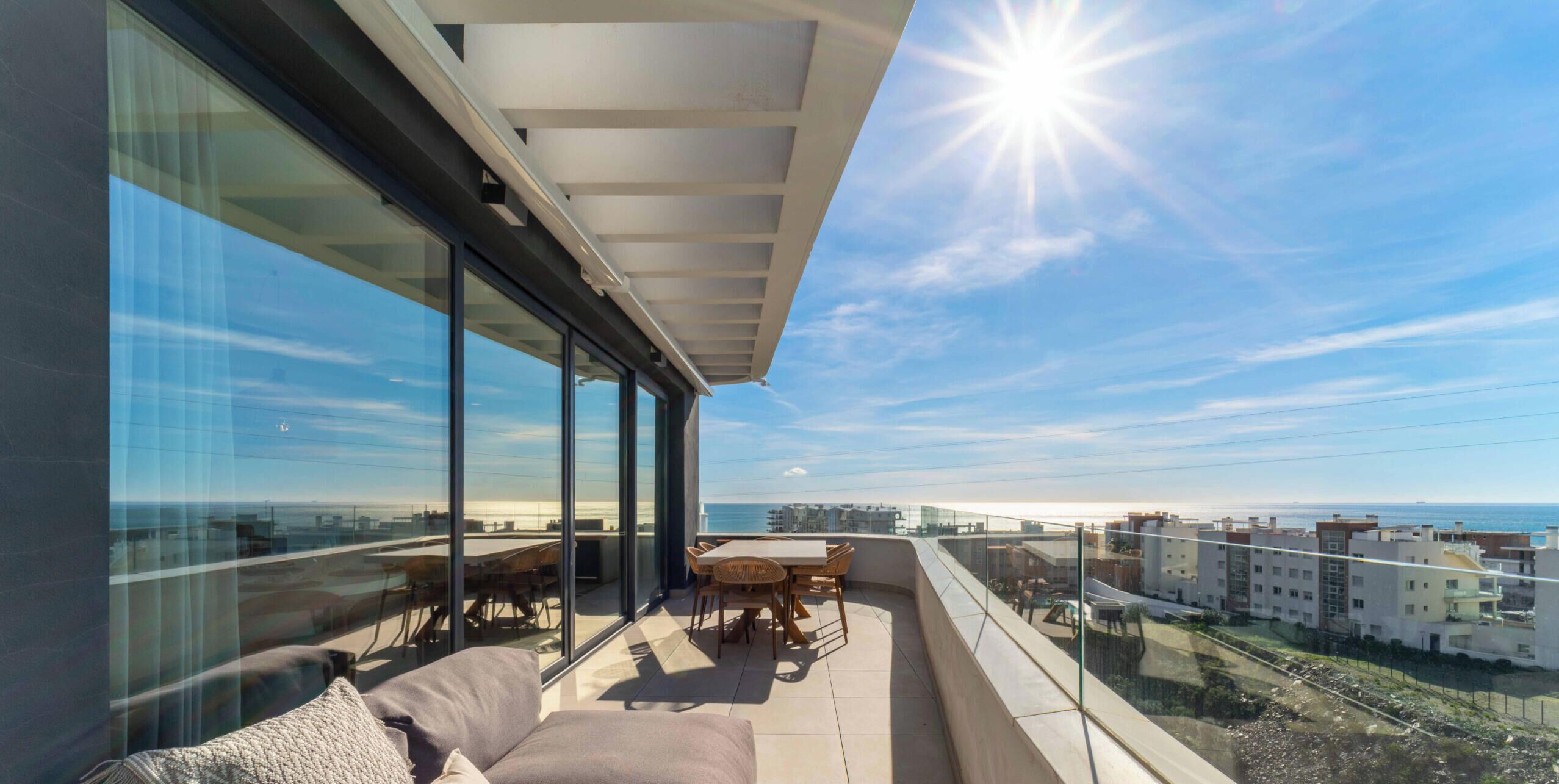 A balcony with a sofa and table, large windows, and a view of the city and sea.