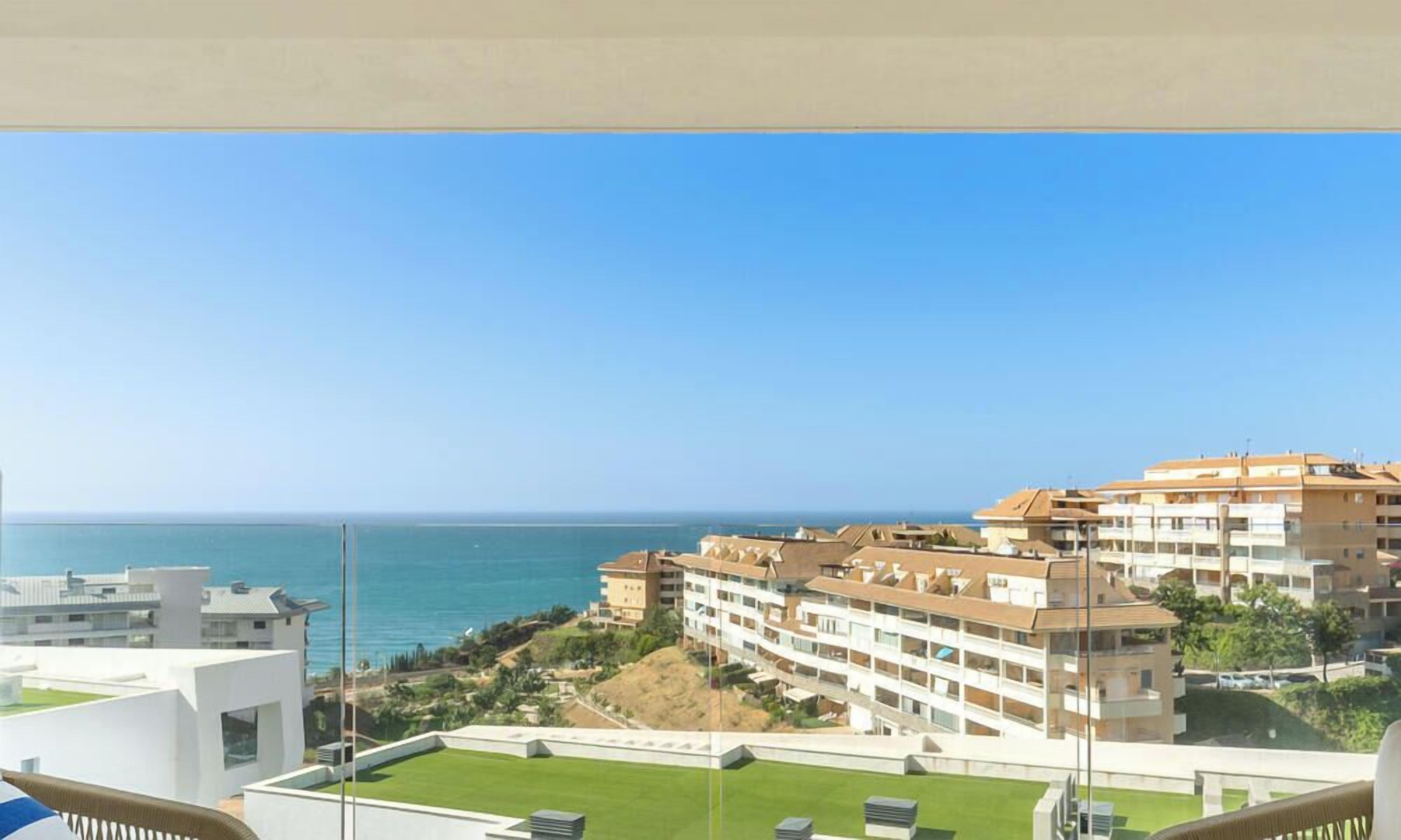 A coastal hotel with multiple balconies overlooking the sea, a well-maintained lawn, and a clear sky.