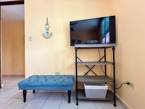A room with a television on a stand, a blue cushioned bench, and a wall-mounted decorative anchor. - 3-bedroom condo in beach town of Rincón with pool and AC at Sea Beach Village (Rincón)