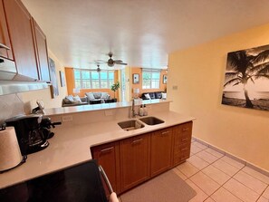 A kitchen with a central island, wooden cabinets, a ceiling fan, and a view of the living area through large windows. - 3-bedroom condo in beach town of Rincón with pool and AC at Sea Beach Village (Rincón)