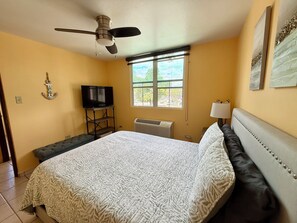 A bedroom with a bed, a television on a stand, a ceiling fan, a window with a view, and a wall-mounted air conditioner. - 3-bedroom condo in beach town of Rincón with pool and AC at Sea Beach Village (Rincón)