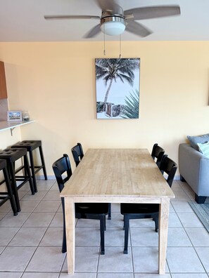 A dining area with a wooden table, black chairs, a ceiling fan, and a wall-mounted picture of a palm tree. - 3-bedroom condo in beach town of Rincón with pool and AC at Sea Beach Village (Rincón)