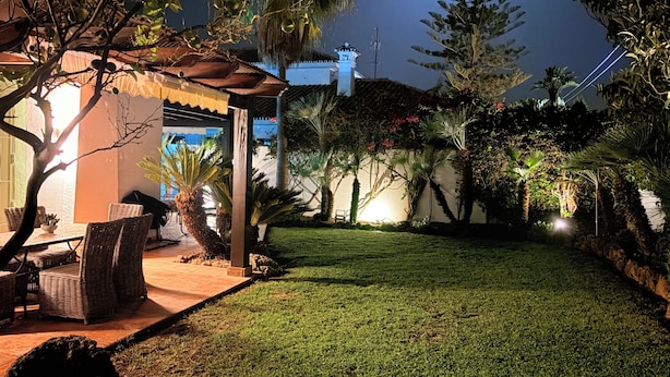 Un espacio exterior bien iluminado con césped, una zona de estar con mobiliario de mimbre y una variedad de plantas y árboles.