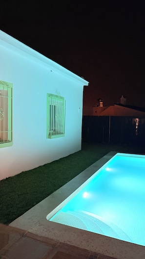 Ein Schwimmbecken mit beleuchtetem Rand, ein weißes Gebäude mit Fenster und eine Rasenfläche.