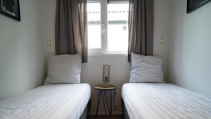 Una habitación pequeña y limpia con dos camas individuales, una mesita y una ventana con cortinas.