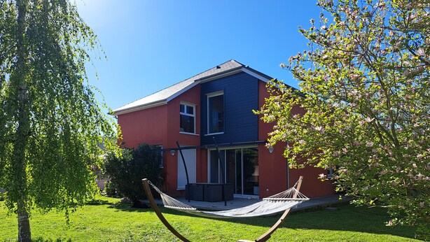 A two-story house with a red exterior and a blue roof, a hammock in the front yard, and a tree with green leaves. - 140 m2 fenced house (Pouzac)