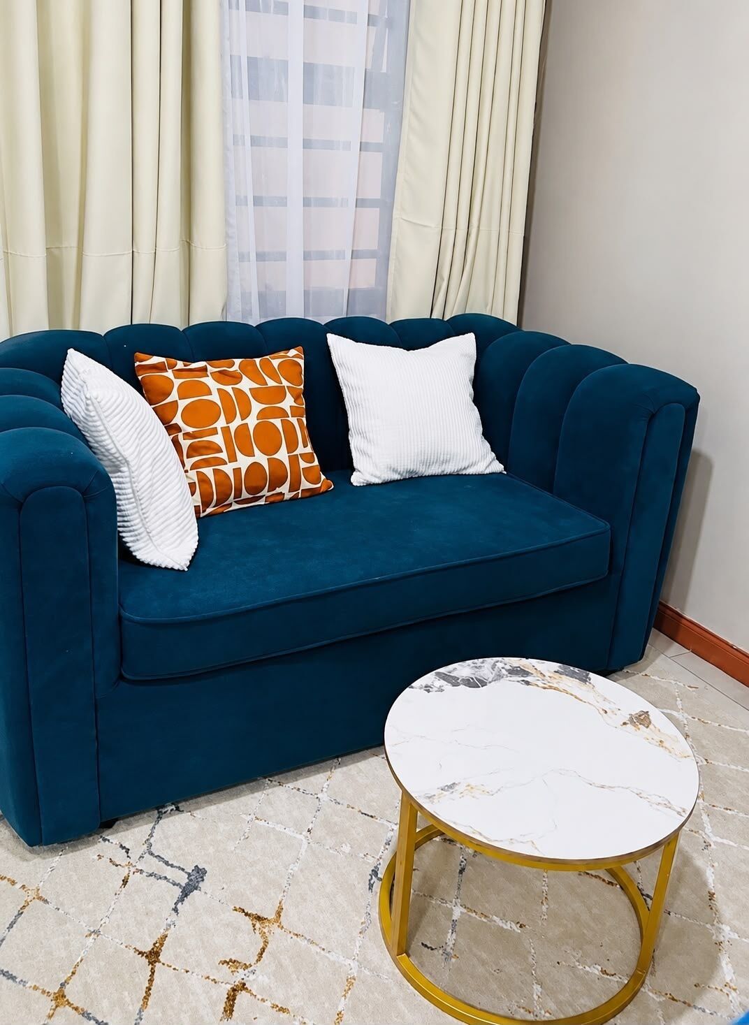 Ein blaues Sofa mit zwei weißen und einem orangefarbenen Kissen, ein Couchtisch aus weißem Marmor mit goldener Basis und ein gemustertes Teppich.