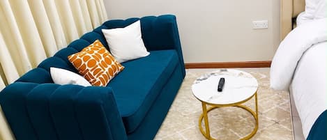 Une pièce avec un canapé bleu, un coussin blanc, un coussin à motifs et une petite table ronde sur laquelle se trouve une télécommande.