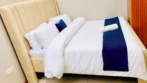 A neatly made bed with white and blue bedding, a beige headboard, and a wall-mounted light fixture. - Veena Homes | Studio Airbnb in Nairobi (Nairobi)