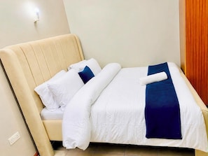 A neatly made bed with white and blue bedding, a beige headboard, and a wall-mounted light fixture. - Veena Homes | Studio Airbnb in Nairobi (Nairobi)