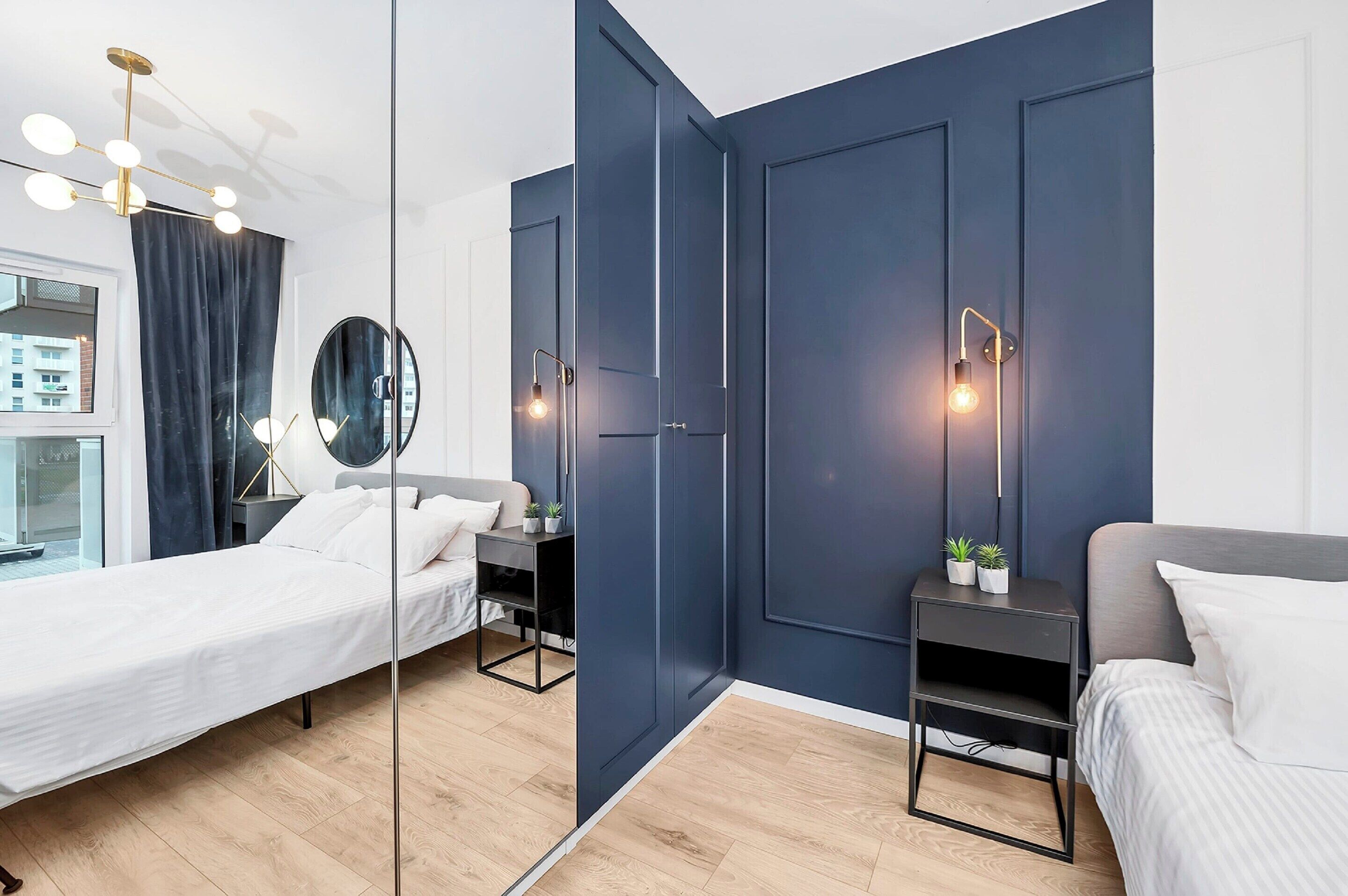 Un dormitorio moderno con un espejo grande, dos camas, mesitas de noche con lámparas y vistas al exterior.