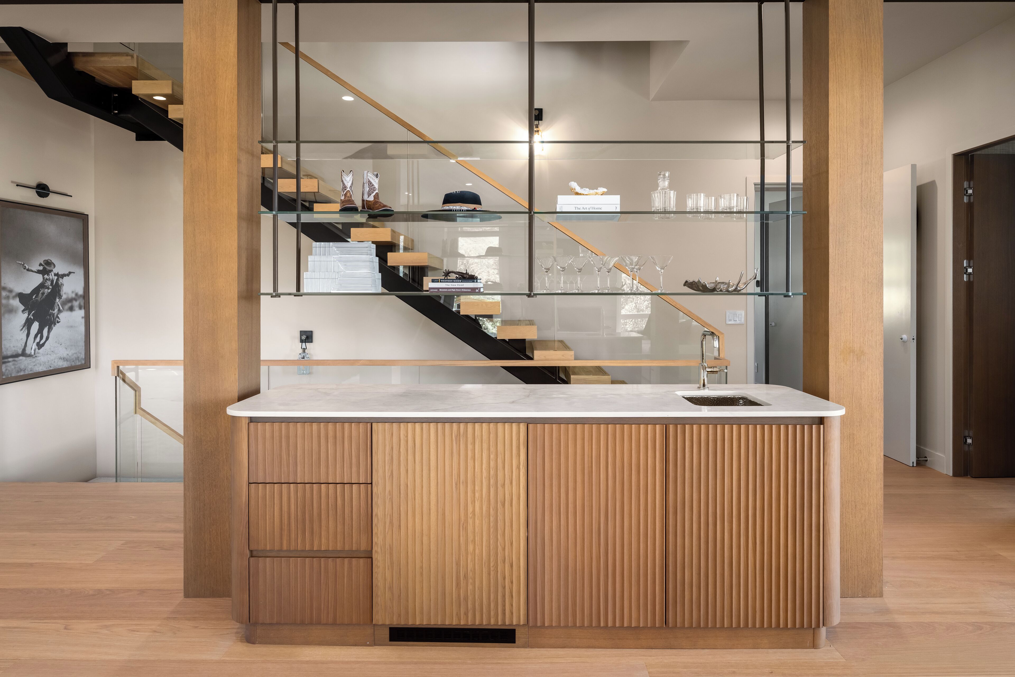 A modern kitchen with a built-in island, wooden cabinetry, and a glass-fronted staircase.