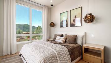 A bedroom with a large window, a bed with a white quilt, two wall-mounted lamps, and two framed pictures.