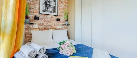 A hotel room with a brick wall, a framed picture, a bed with rolled towels, a bouquet of flowers, and a magazine.