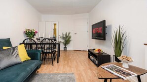 Una sala de estar con un sofá, una mesa de comedor con sillas, un televisor montado en la pared y una estantería.