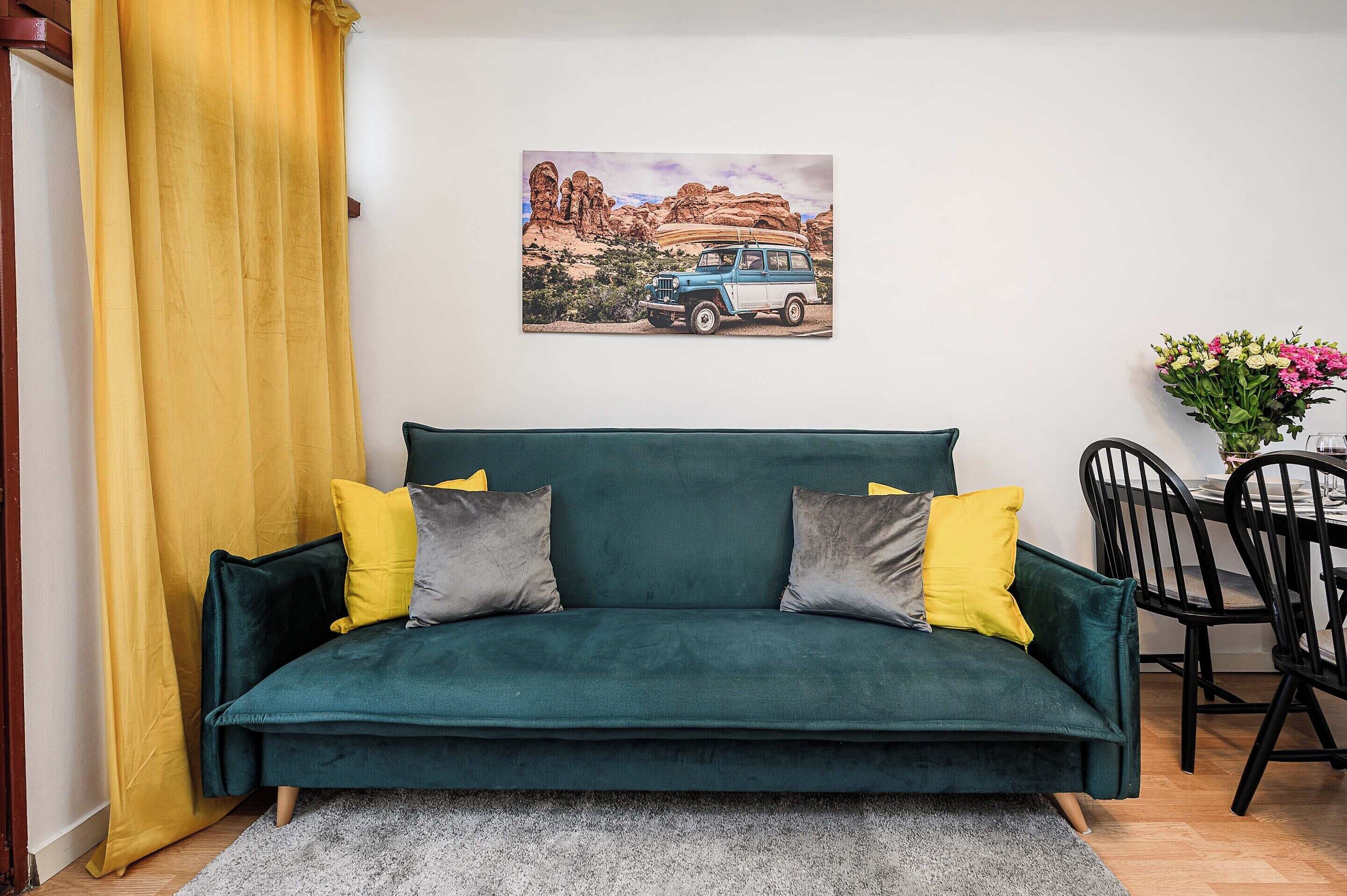 Uma sala de estar aconchegante com um sofá verde, duas almofadas, um tapete, uma mesa de jantar com cadeiras e um quadro de um veículo na parede.