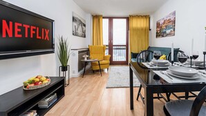 Una sala moderna con un televisor de pantalla plana mostrando 'NETFLIX', un comedor con una mesa dispuesta para dos personas, y un sillón amarillo.