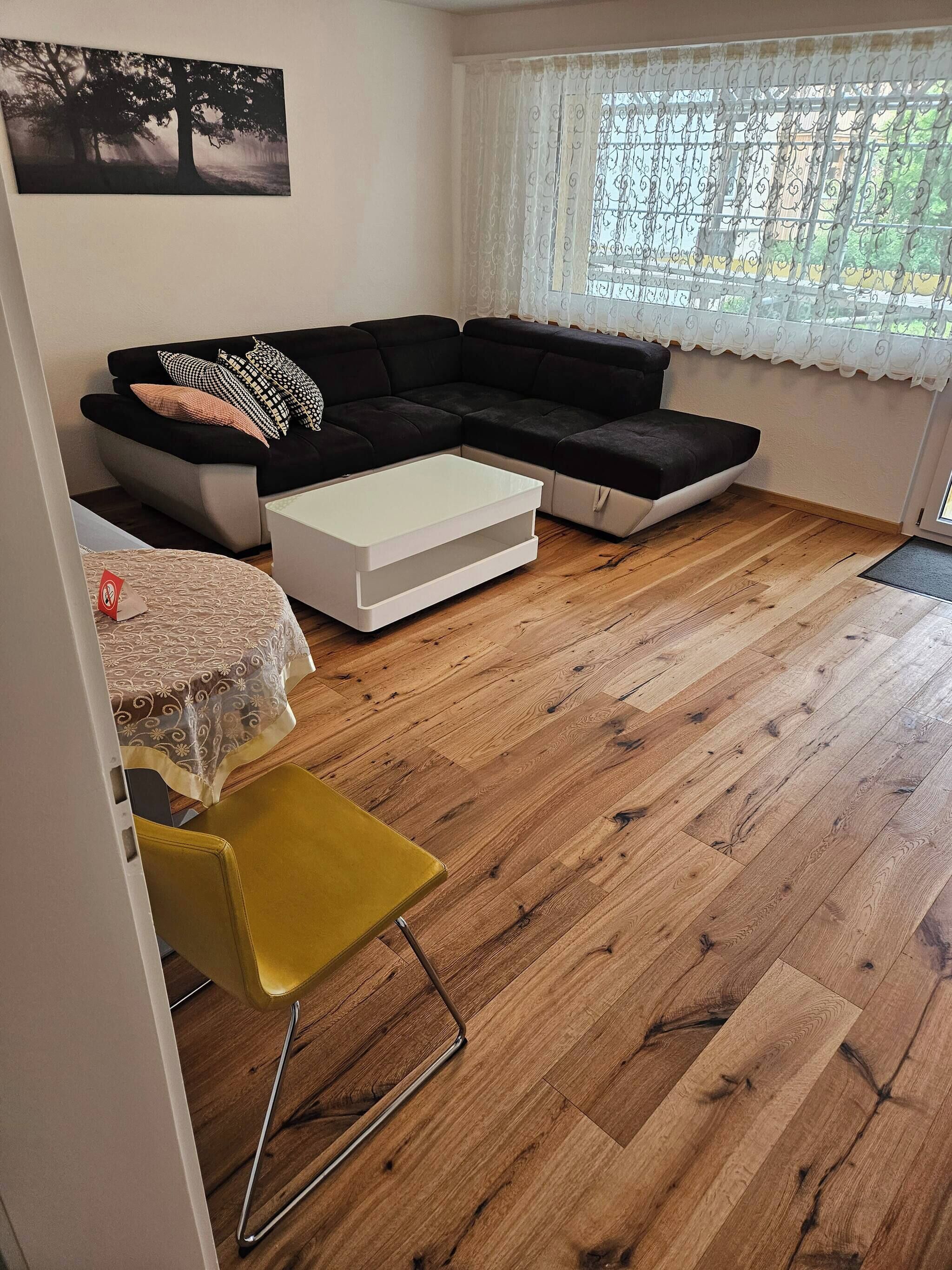 Ein Wohnzimmer mit einem Ecksofa, einem kleinen weißen Couchtisch, einem gelben Sessel und einem Esstisch mit einem gemusterten Tischläufer.