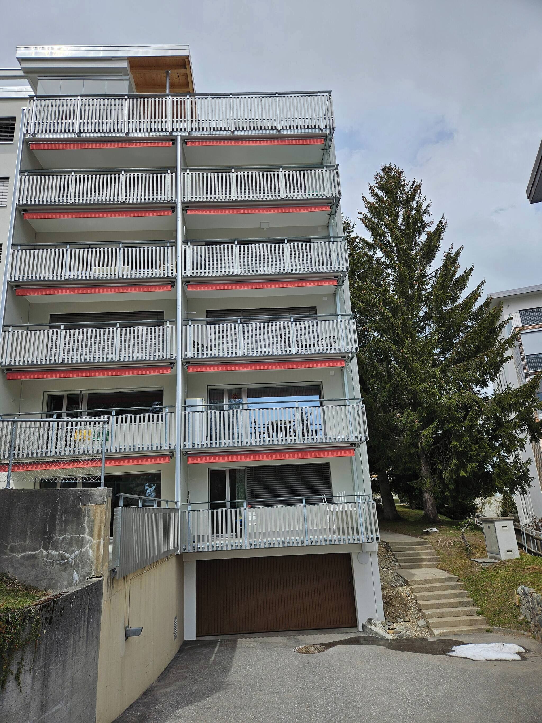 A multi-story residential building with balconies, a garage, and a tree in the background.
