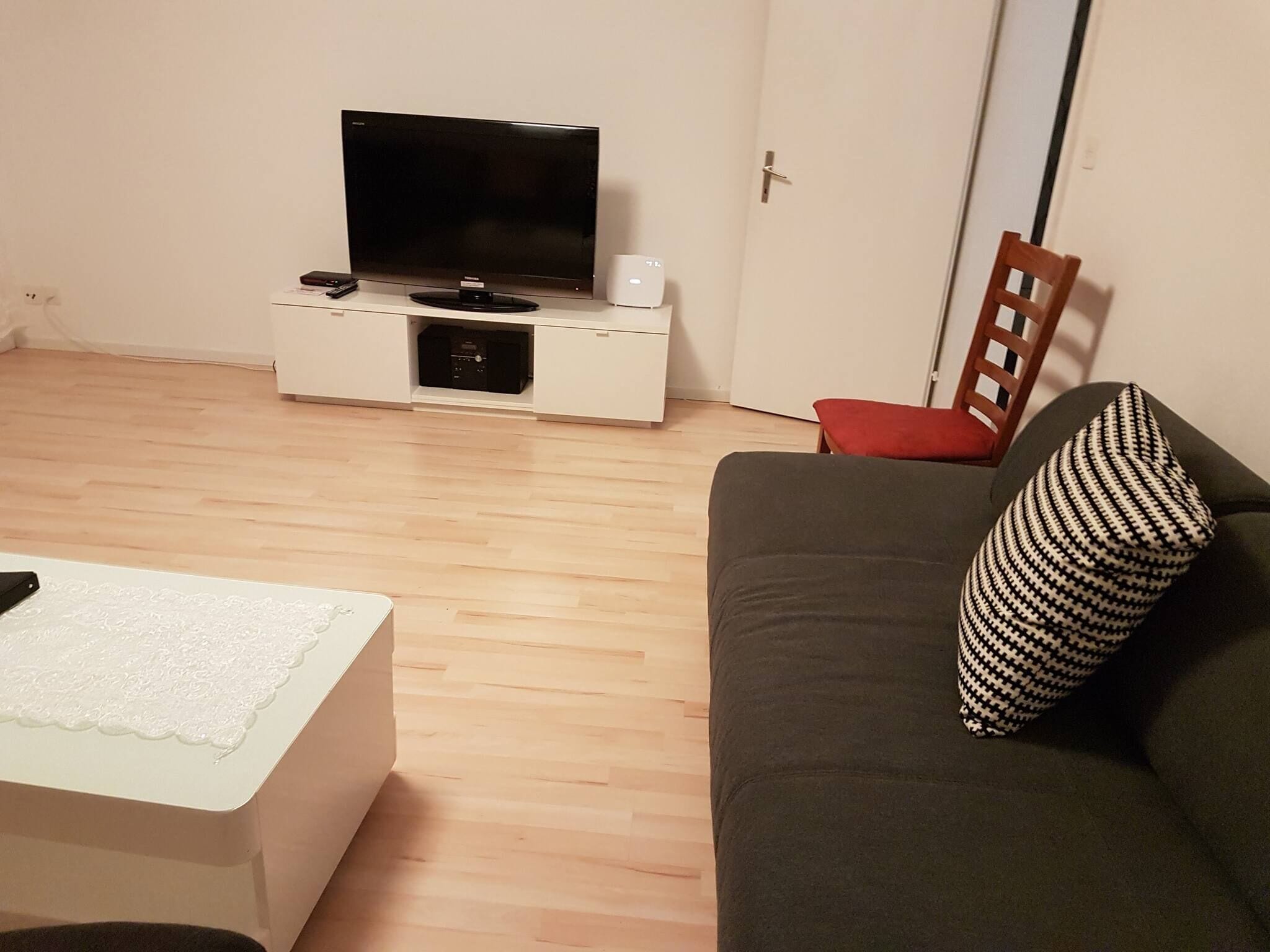 A living room with a flat-screen TV on a white stand, a grey sofa with a patterned pillow, and a red chair.