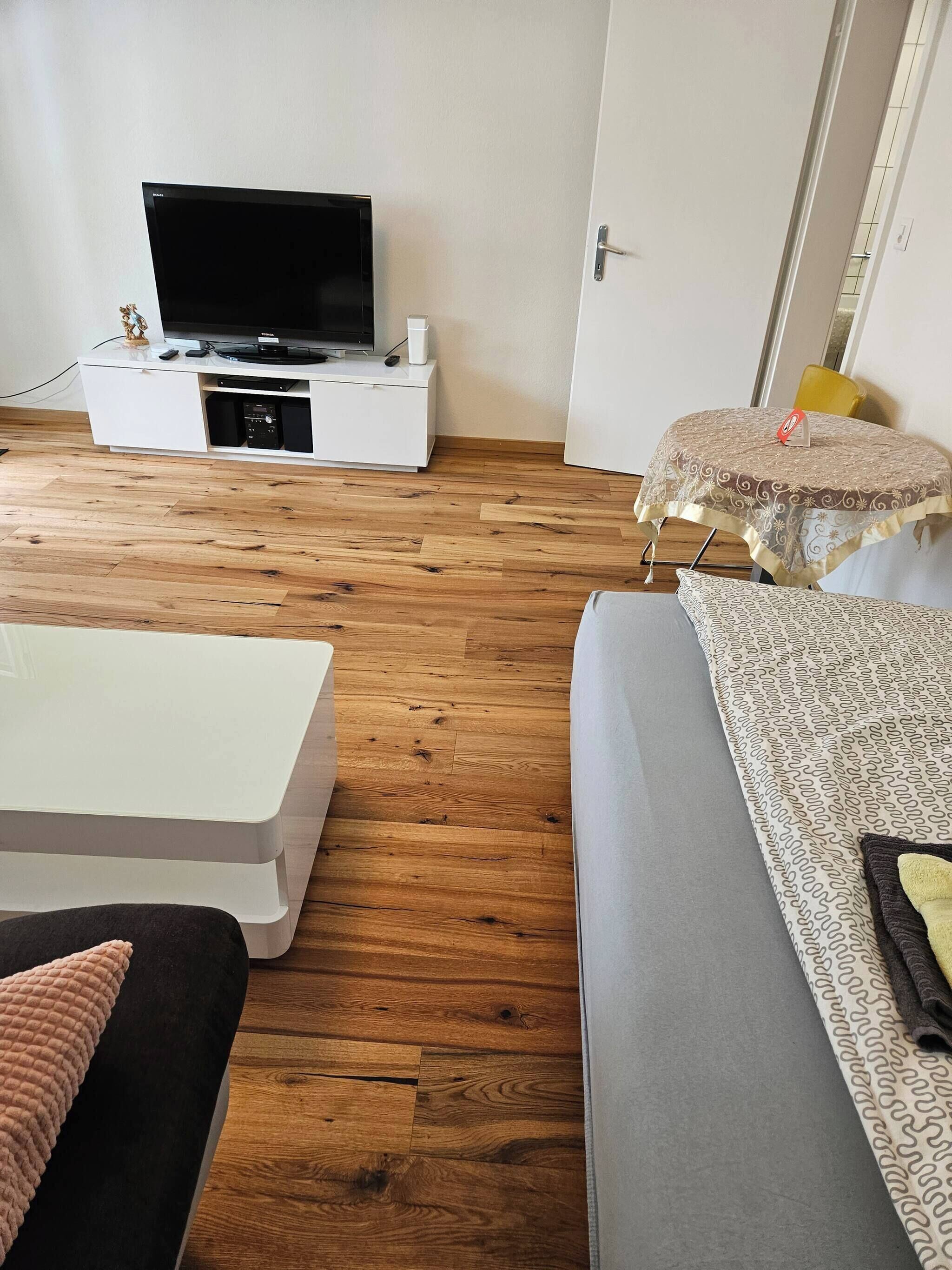 Una sala de estar con un televisor de pantalla plana sobre un soporte blanco, un sofá, una mesa de centro y un mantel estampado sobre una mesita redonda.