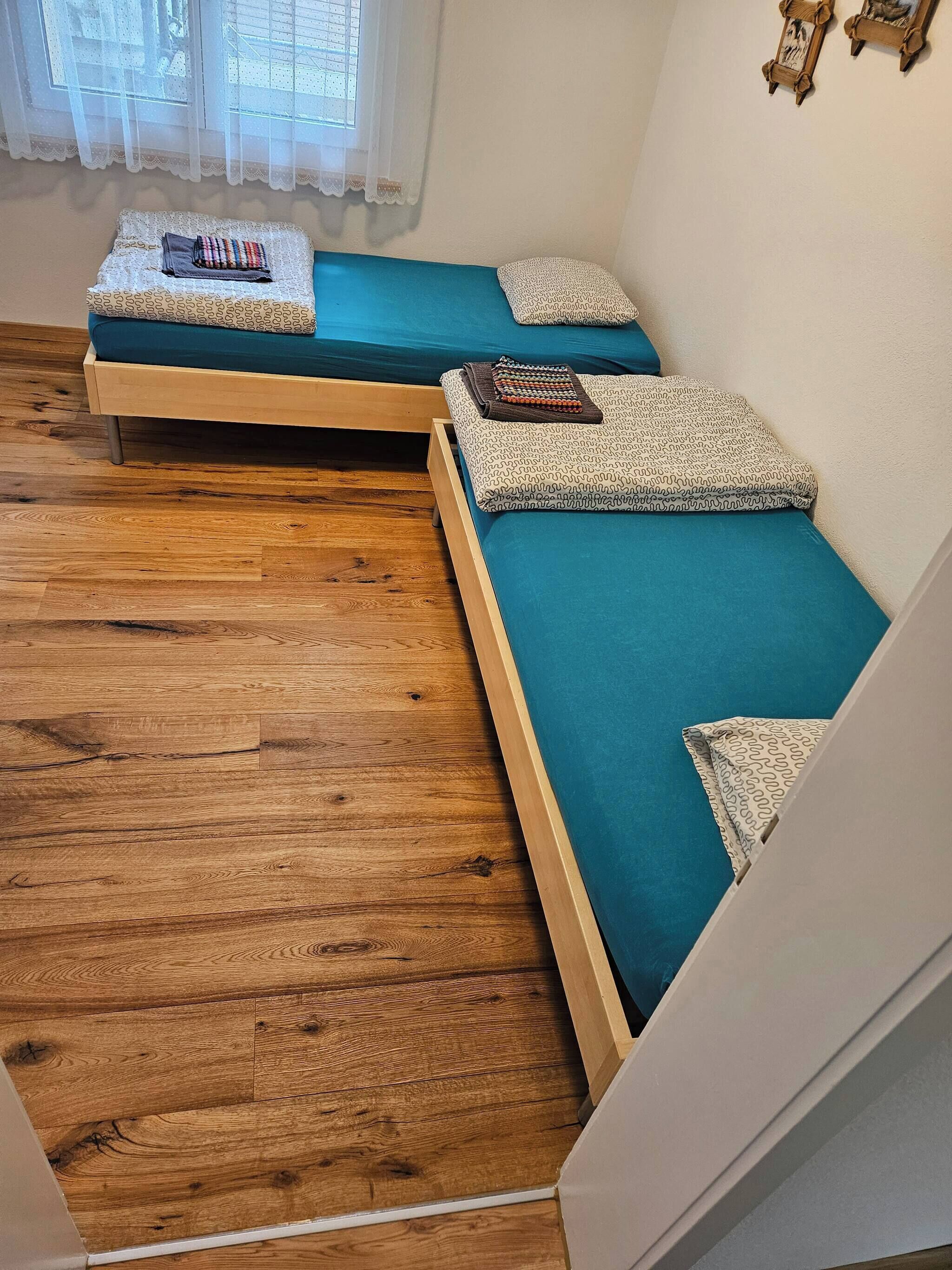 Ein kleines Zimmer mit zwei Einzelbetten, Holzboden und einem Fenster mit durchsichtigen Vorhängen.