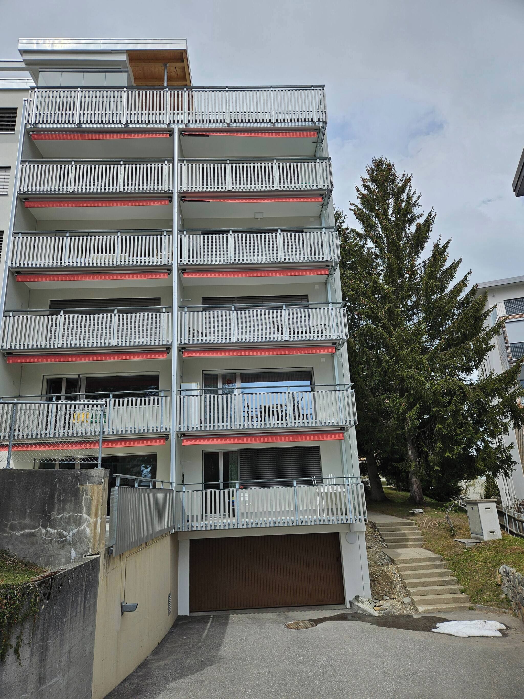 A multi-story residential building with balconies, a garage, and a tree in the background.