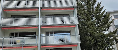 Un edificio residencial de varios pisos con balcones, un garaje y un árbol al fondo.
