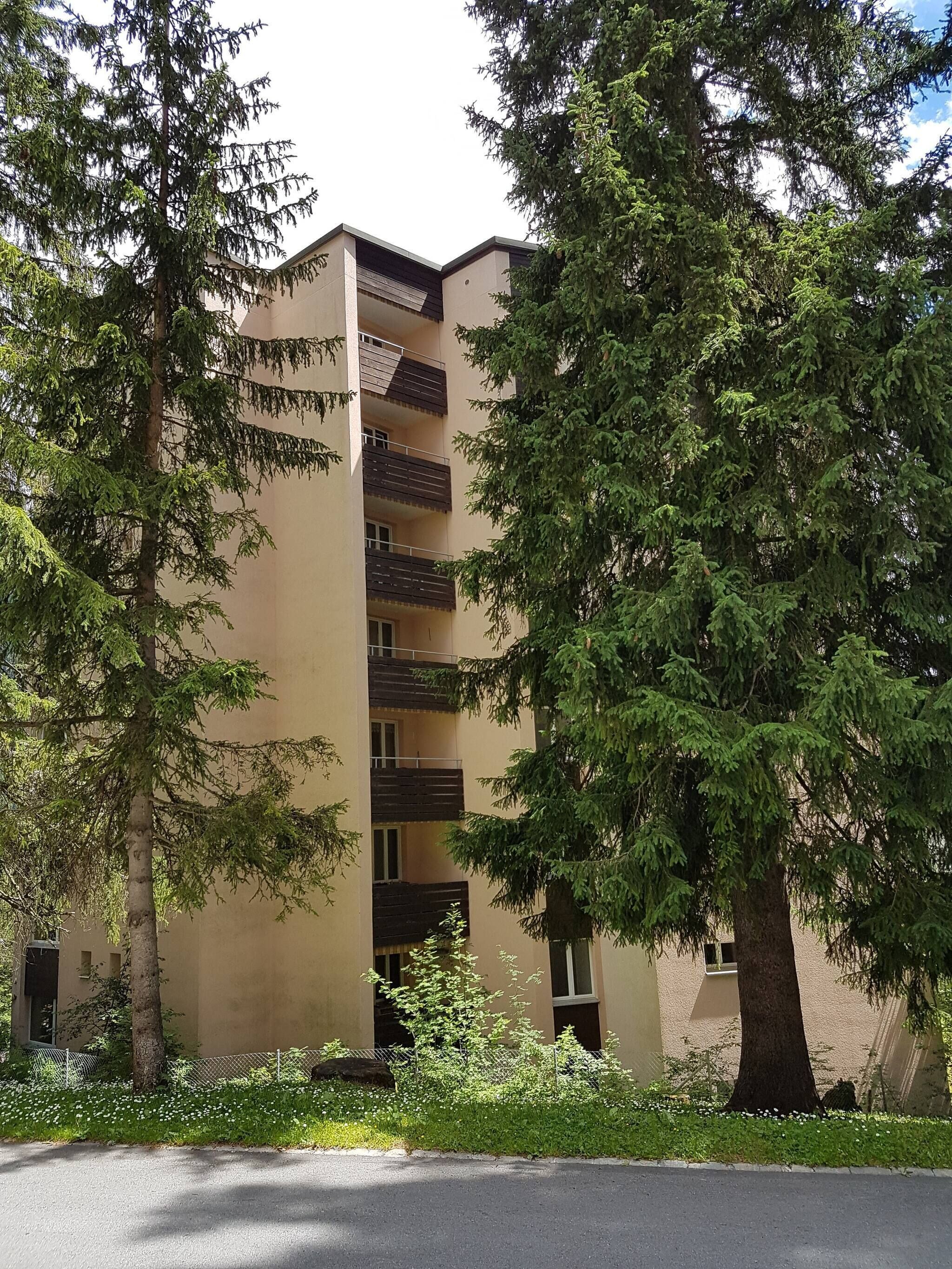 A multi-story building with balconies, surrounded by trees and a grassy area.