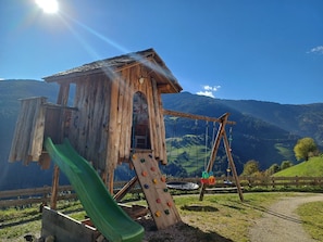 A wooden play structure with a slide and climbing wall, set against a mountainous backdrop.