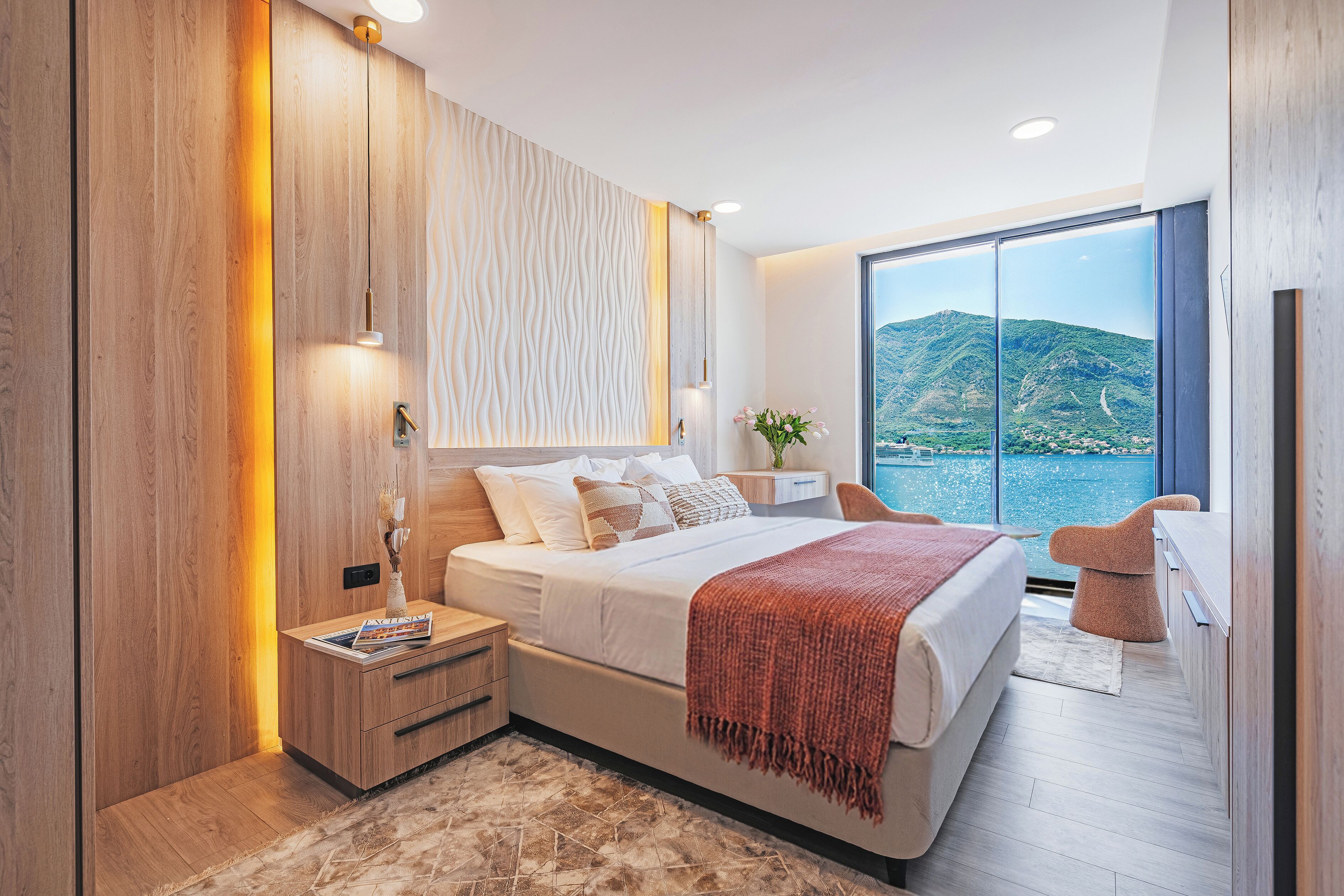 A modern bedroom with a large bed, bedside tables, a nightstand, a window with a view of a mountain and water, and a wooden wall.