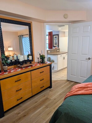 Ein Schlafzimmer mit einem hölzernen Schminktisch, einem großen Spiegel, einem Bett mit grüner Decke und einer Tür zu einem Badezimmer.