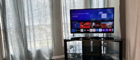 A flat-screen TV on a black stand in a room with a window and curtains.